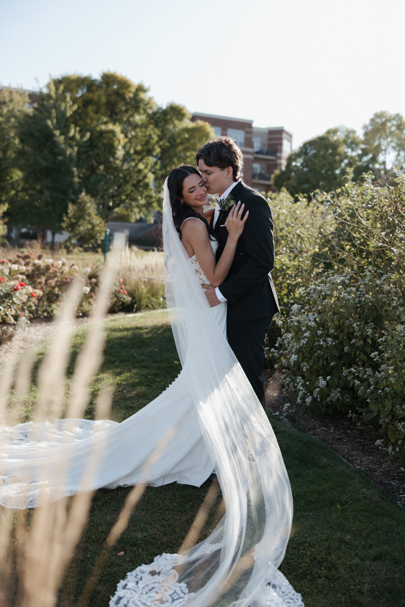 Groom kissing bride on the cheek while her veil flows towards the camera