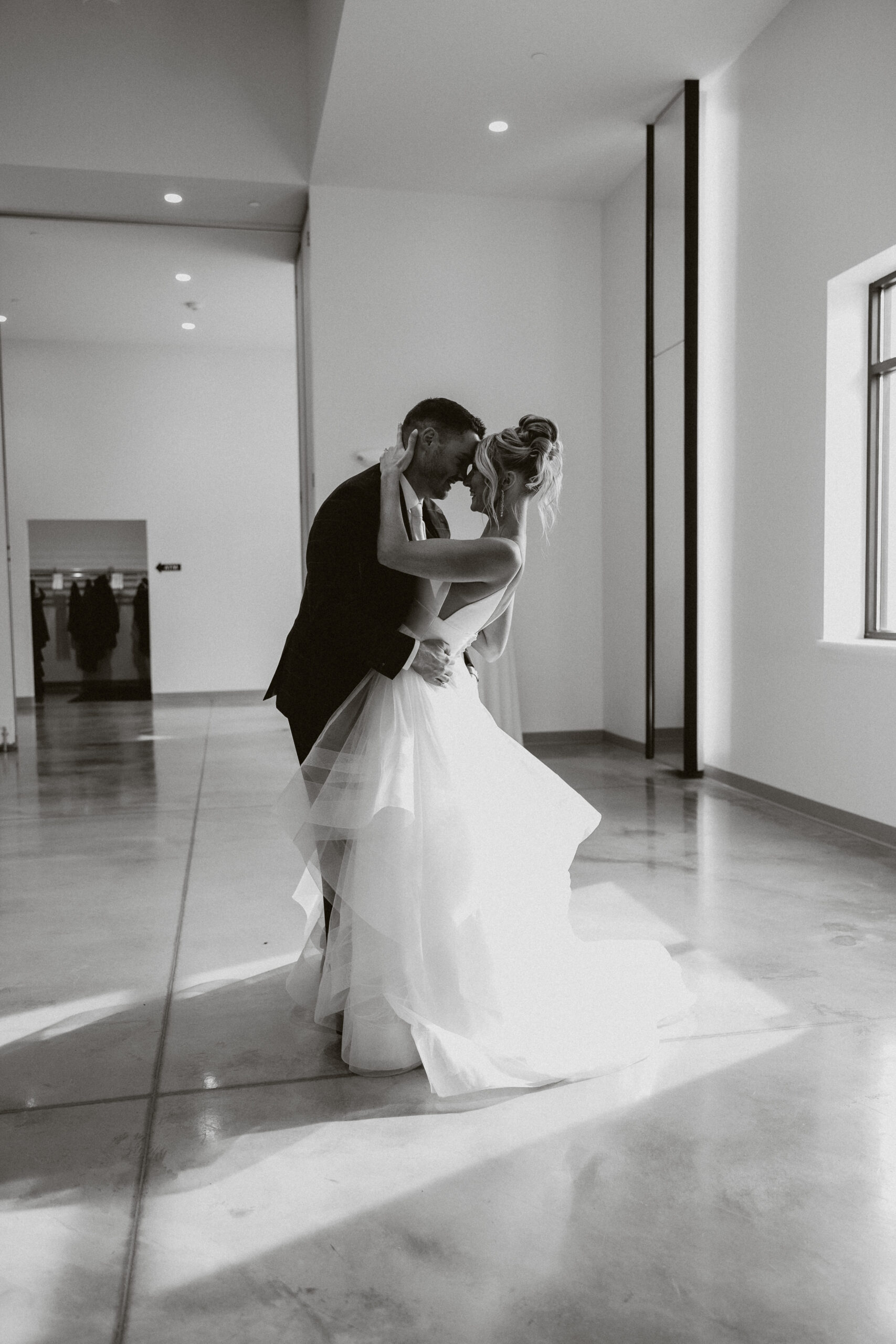 A black and white photo of a bride and groom touching foreheads by a window inside of the Valerie