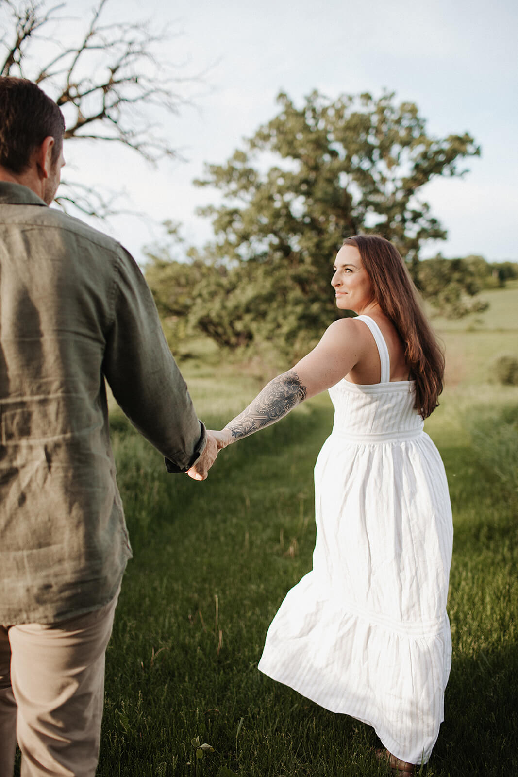 Couple walking hand in hand through a green grassy field with trees and open countryside surrounding them during an outdoor engagement session.