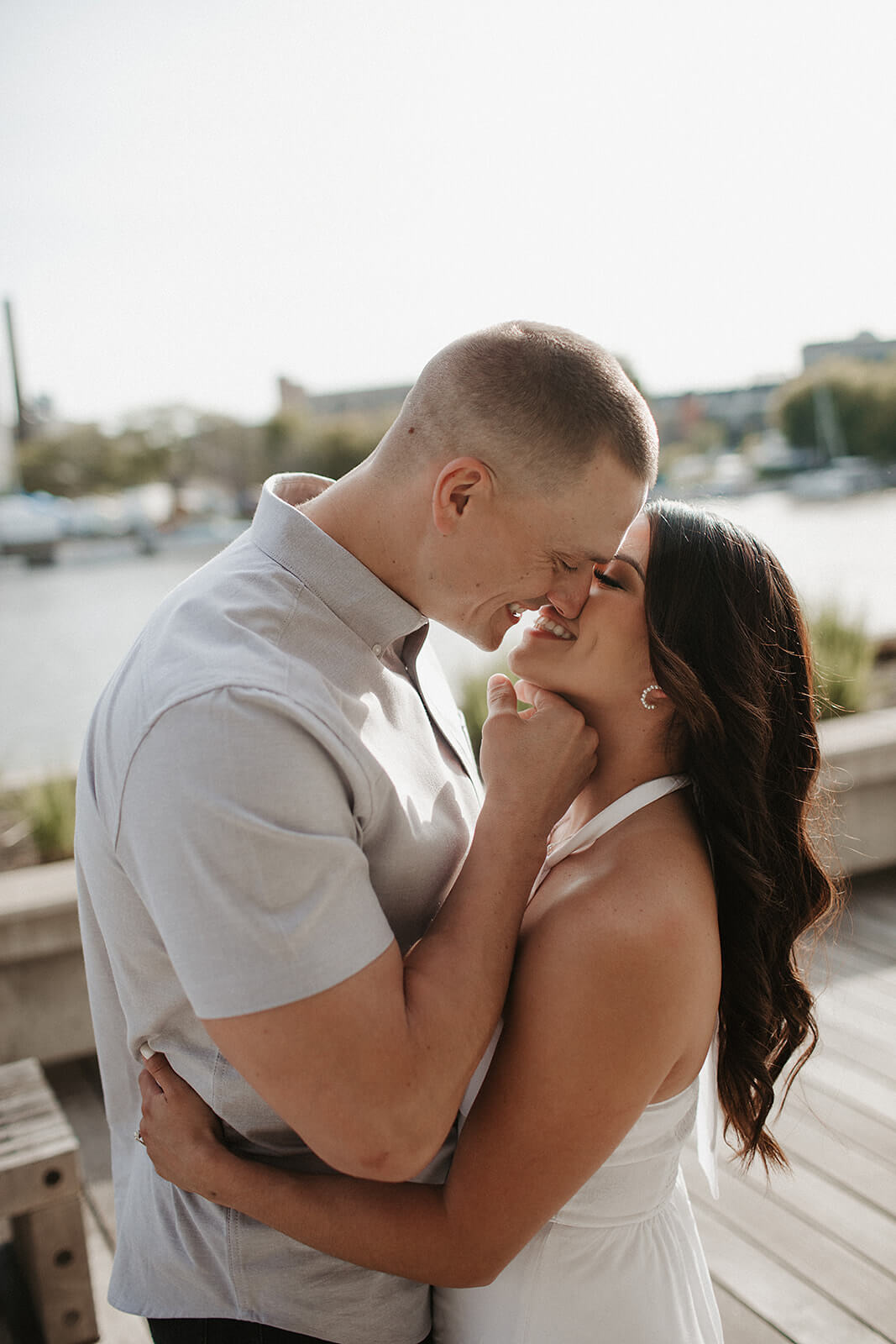 Close-up portrait of a couple embracing on a wooden boardwalk near the water with boats and a marina blurred in the background.