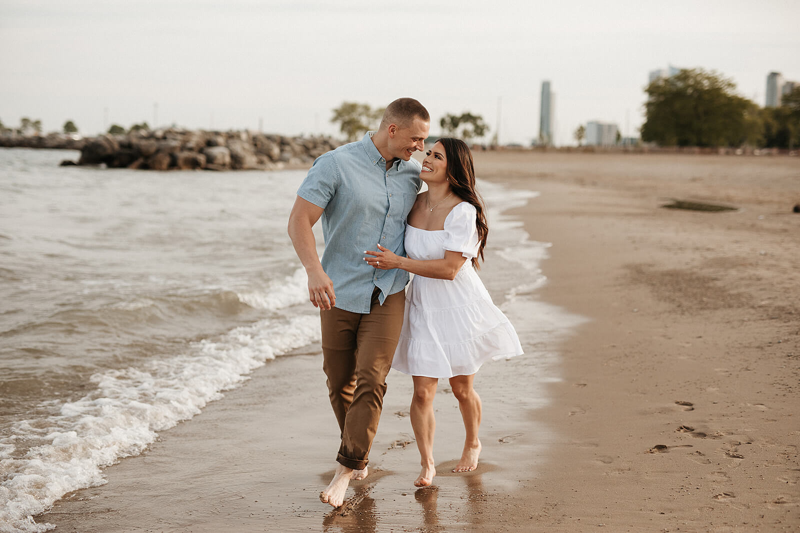 Couple walking barefoot together along the edge of the beach with gentle waves washing onto the sand and a city skyline in the distance.