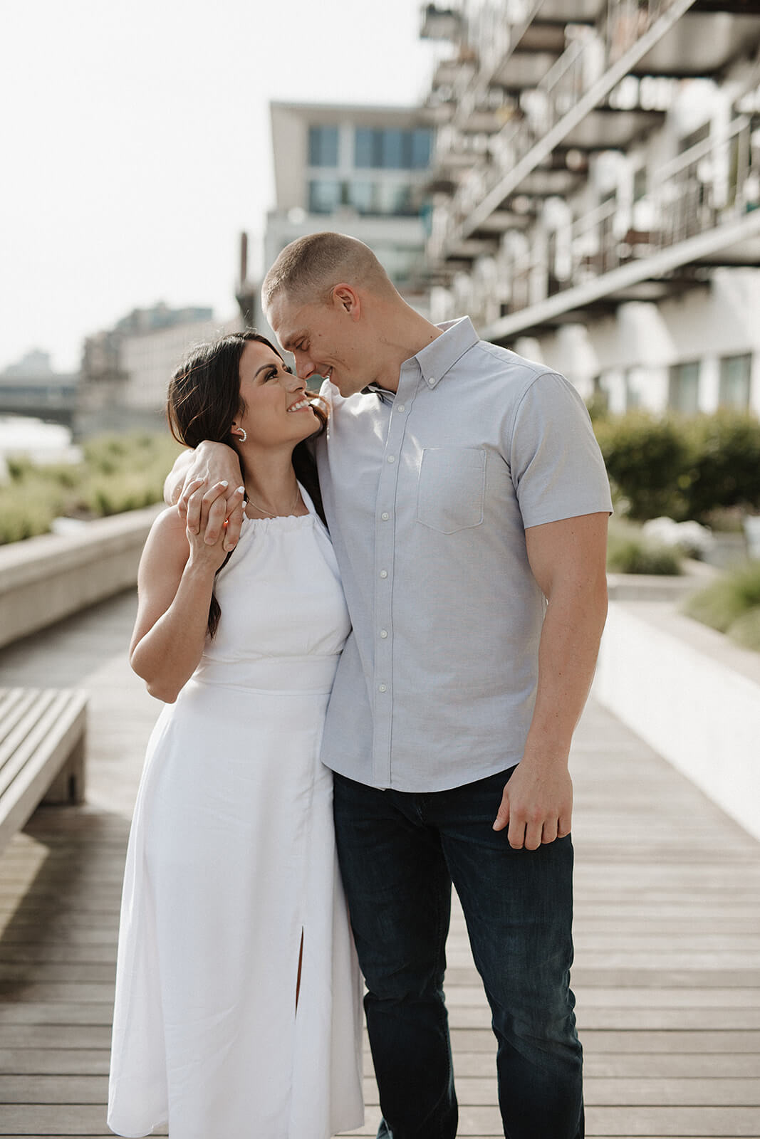 Couple standing together on a wooden boardwalk near beachfront condos, smiling and embracing during a waterfront engagement session.