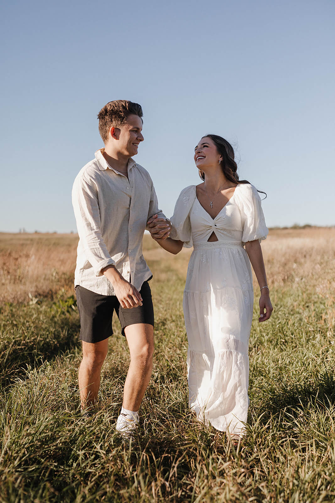 Couple walking together through a grassy field during sunset while smiling at each other and holding hands.