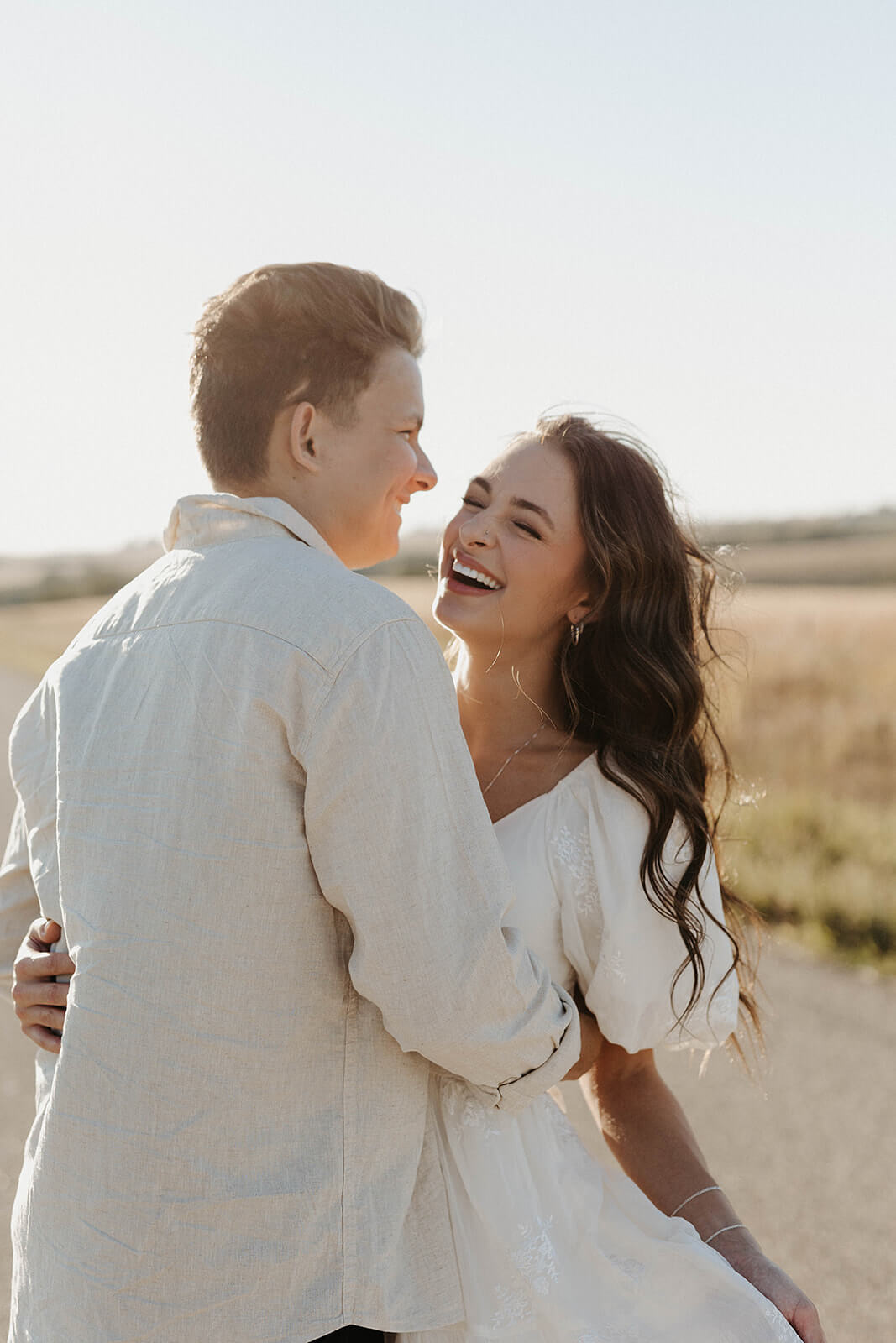 Couple embracing and smiling together on a country road during golden hour with tall grass and open countryside surrounding them.