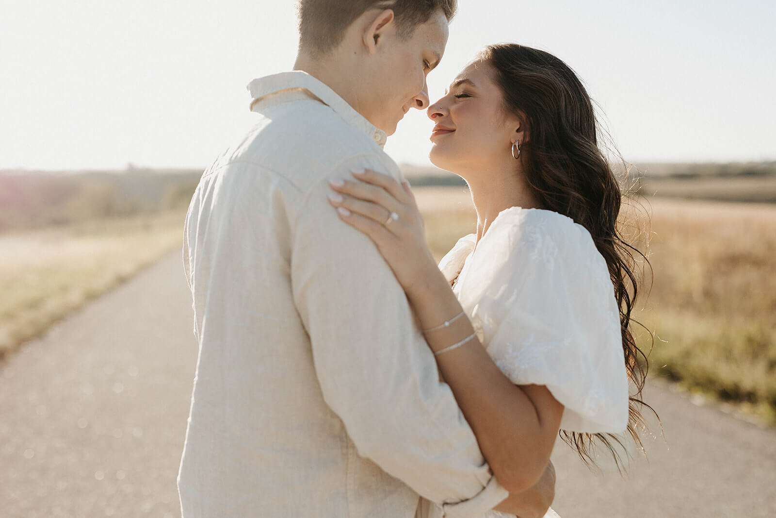 Close-up portrait of a couple embracing on a country road during golden hour, with the woman resting her hand on the man’s shoulder and showing her engagement ring.