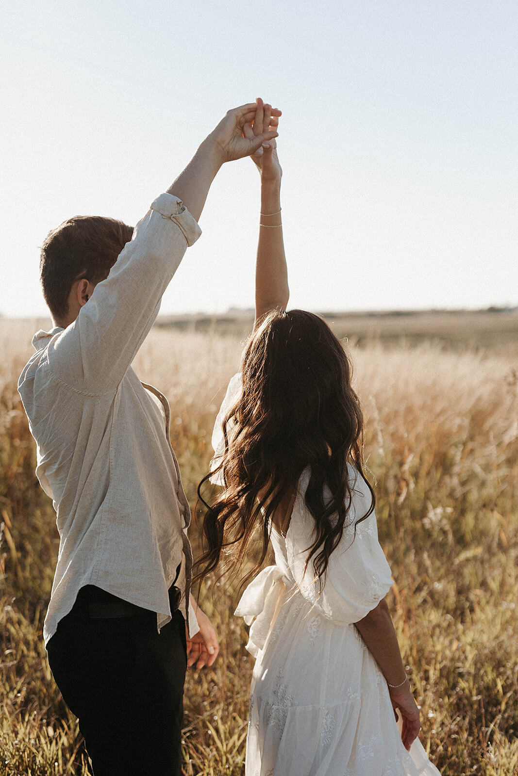 Couple holding hands while twirling together through a golden grassy field during sunset, with soft sunlight illuminating the scene.