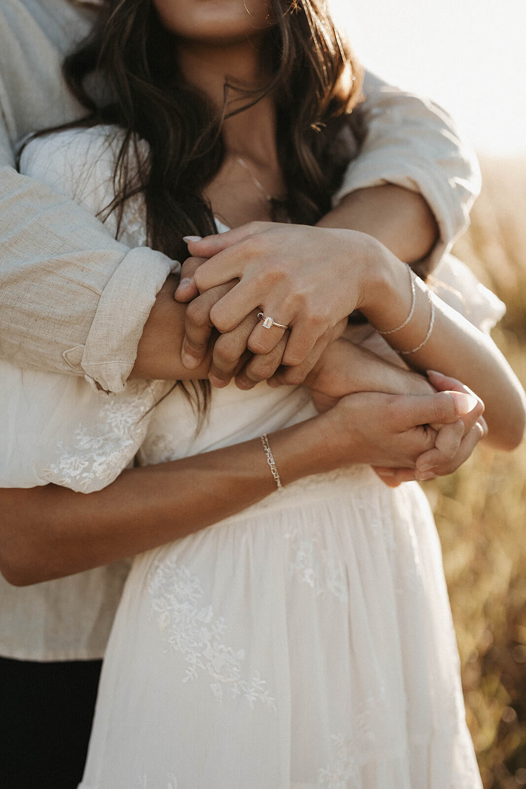 Close-up detail photo of a couple embracing, focused on their hands and engagement ring while standing in a grassy field during golden hour.