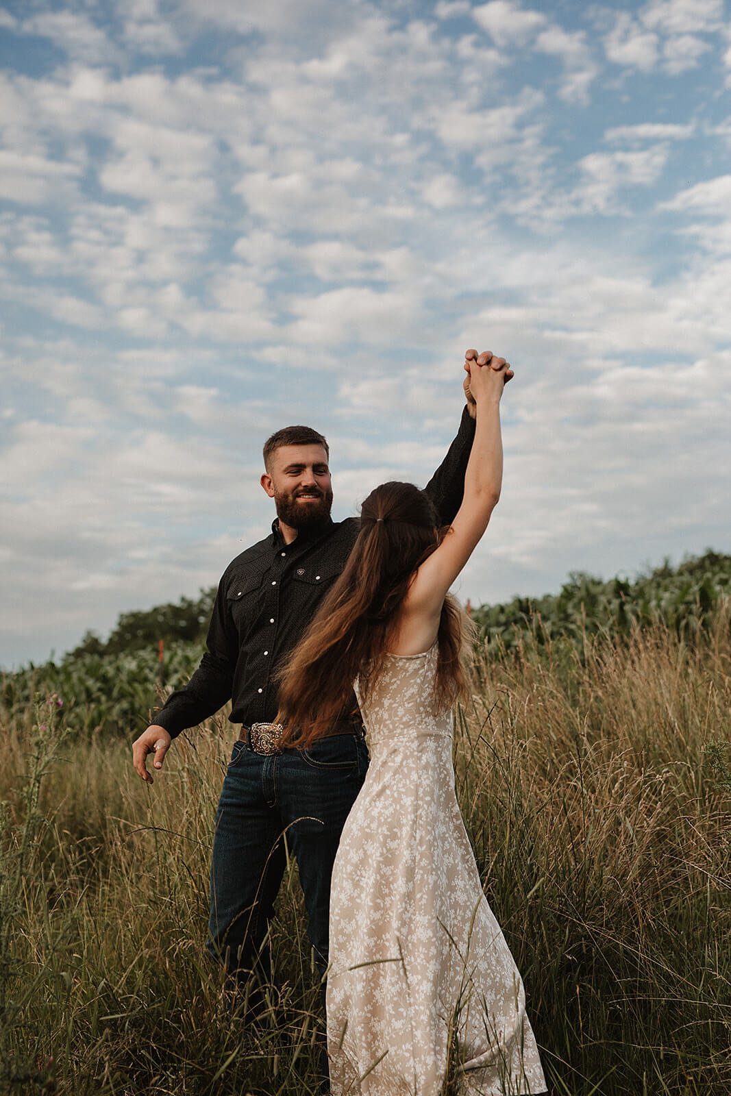 Couple dancing together in a grassy field beneath a blue sky filled with soft clouds while holding hands during golden hour.