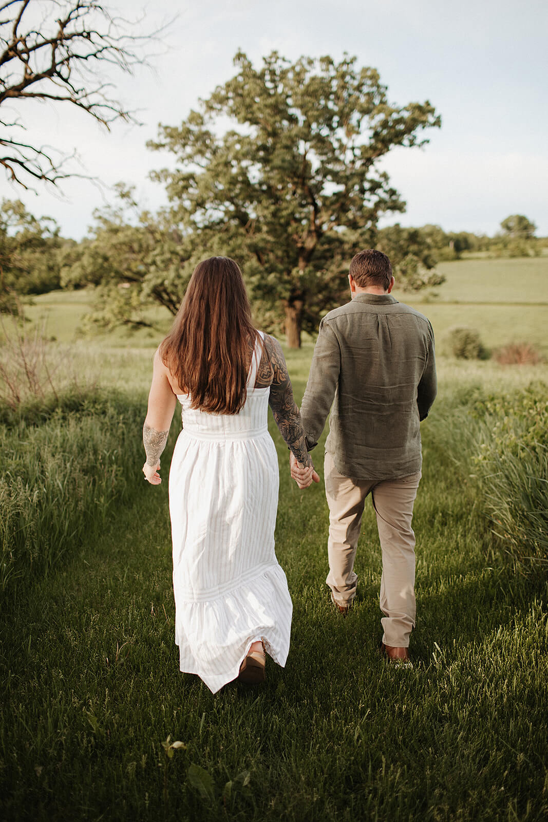Couple walking hand in hand through a grassy field with rolling countryside and trees surrounding them during an outdoor engagement session.