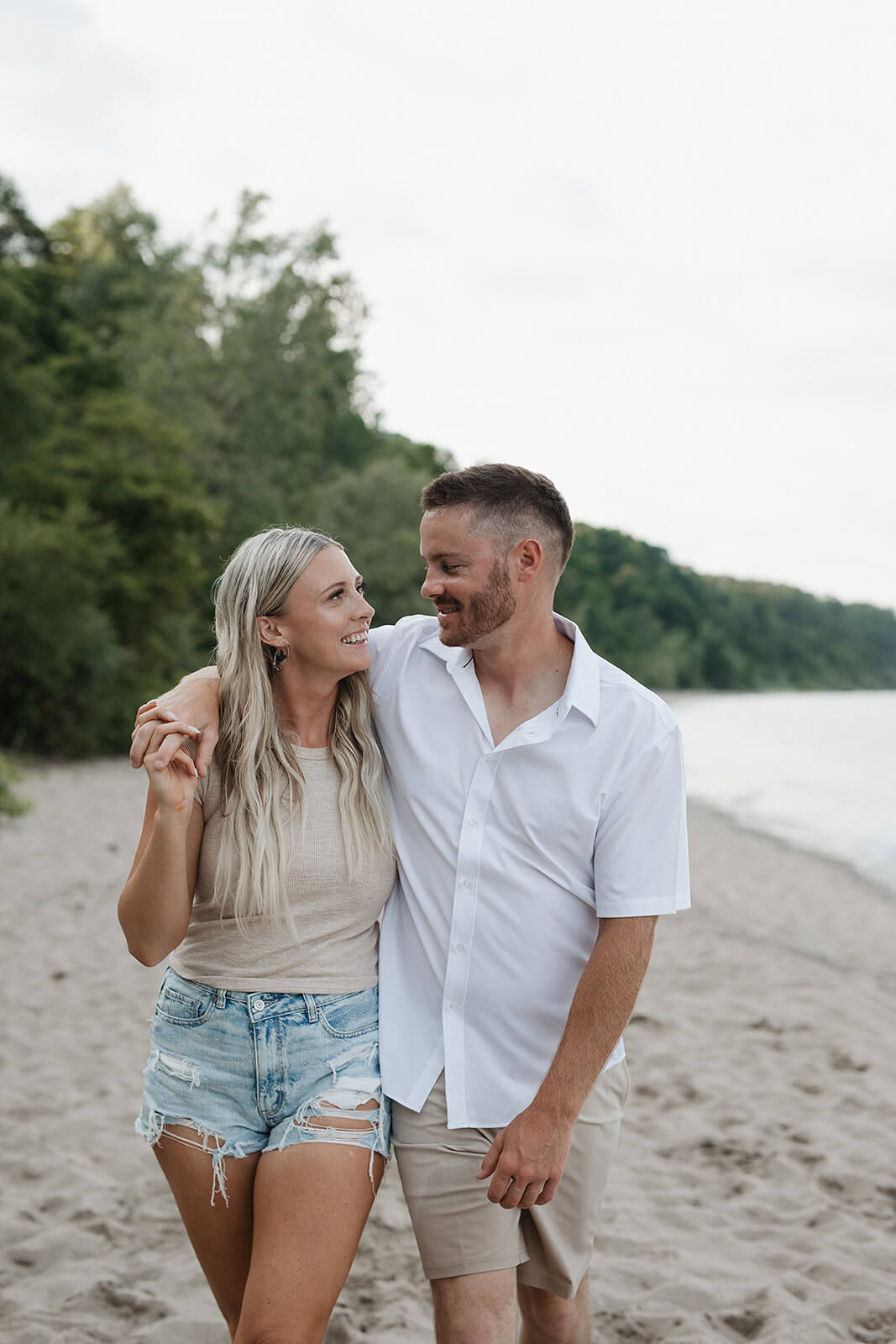 Couple walking together along a sandy beach shoreline while smiling at each other during an outdoor engagement session.