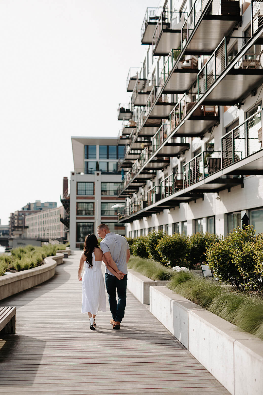 Couple walking together on a waterfront boardwalk lined with modern apartment buildings and greenery during an urban engagement session.