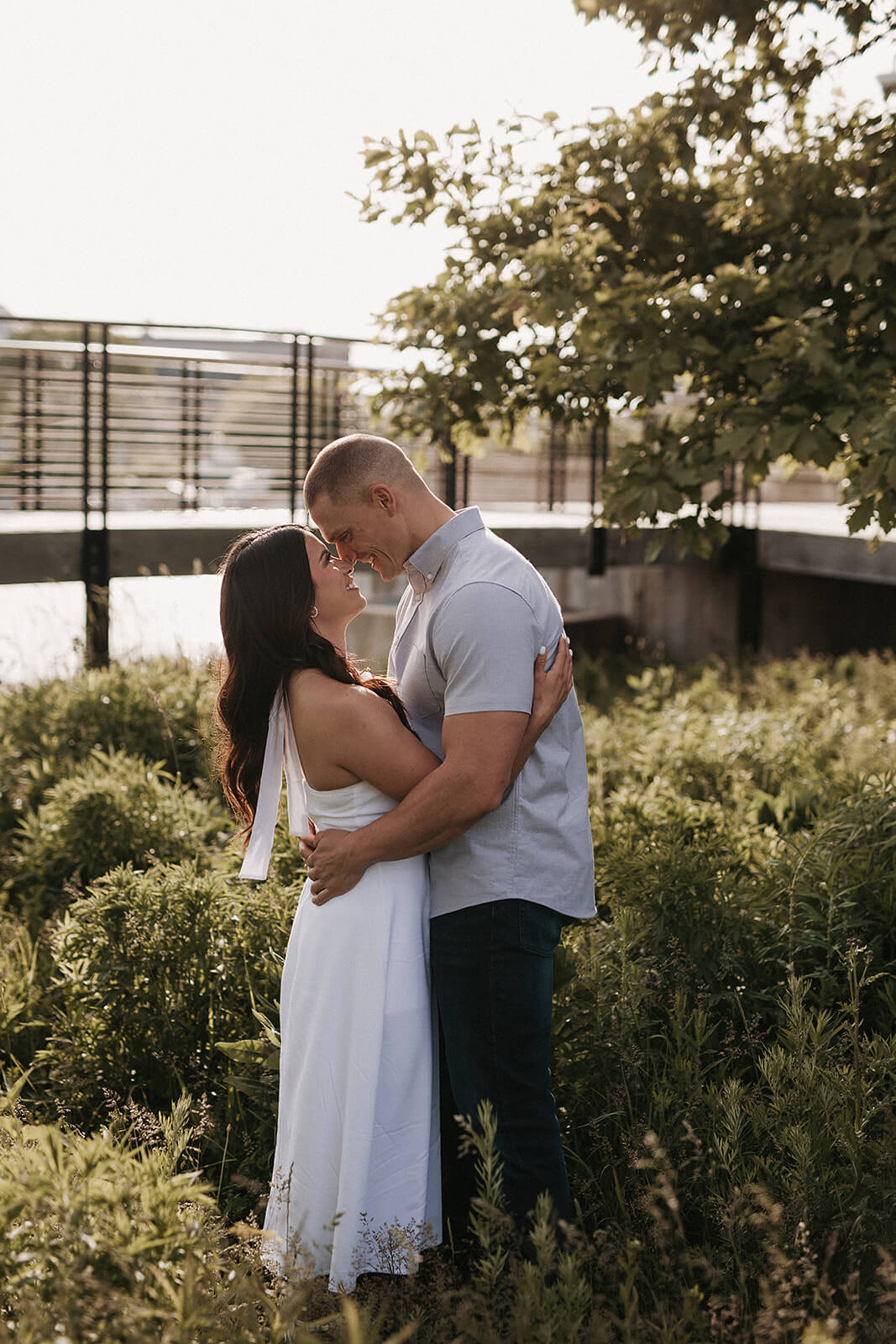 Couple embracing outdoors near a waterfront garden with greenery and modern buildings softly blurred in the background.