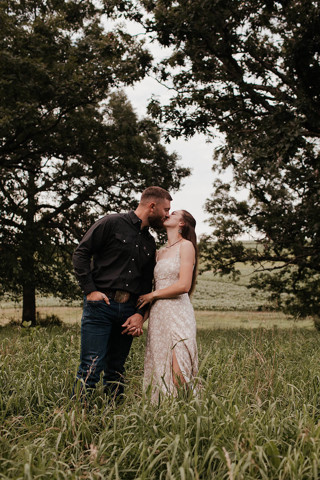 Couple standing together in tall grass beneath large trees during golden hour while embracing in an open field.