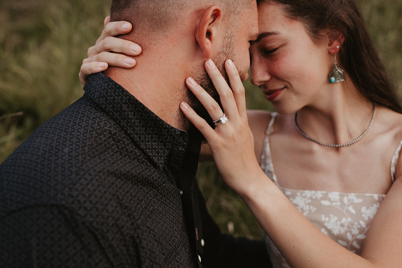 Close-up portrait of a couple embracing outdoors in a grassy field, with the woman showing her engagement ring while resting her hand on the man’s neck.