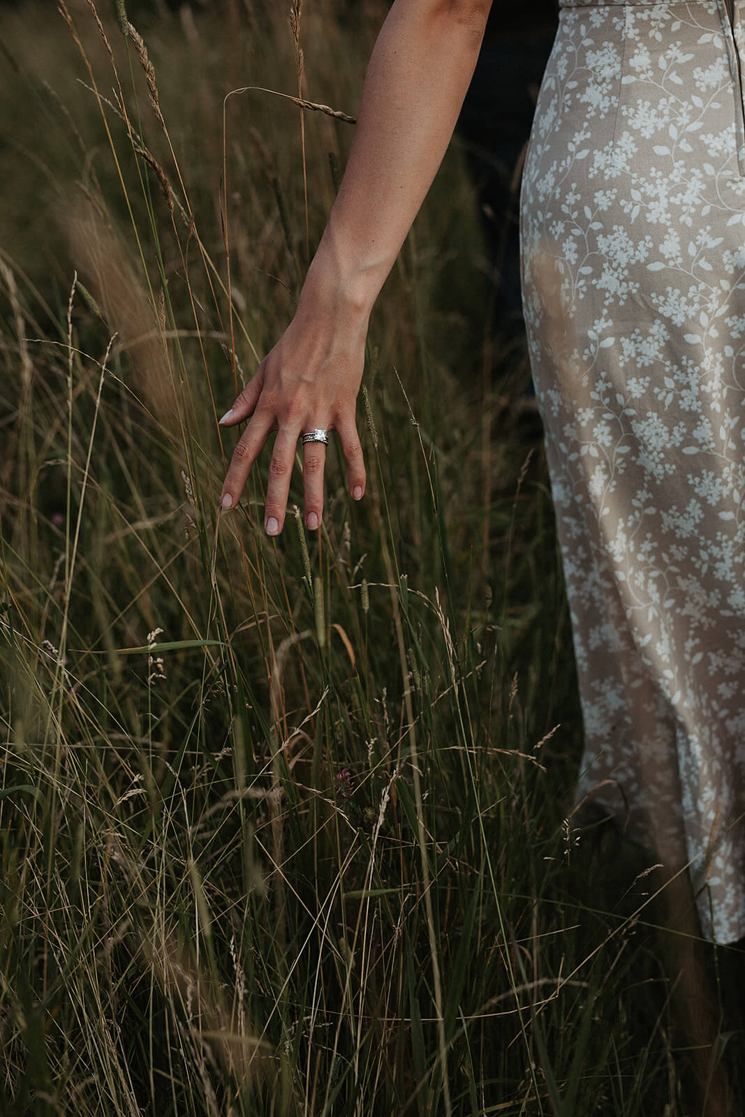 Close-up photo of a woman’s hand brushing through tall grass, showing her engagement ring and floral dress details.