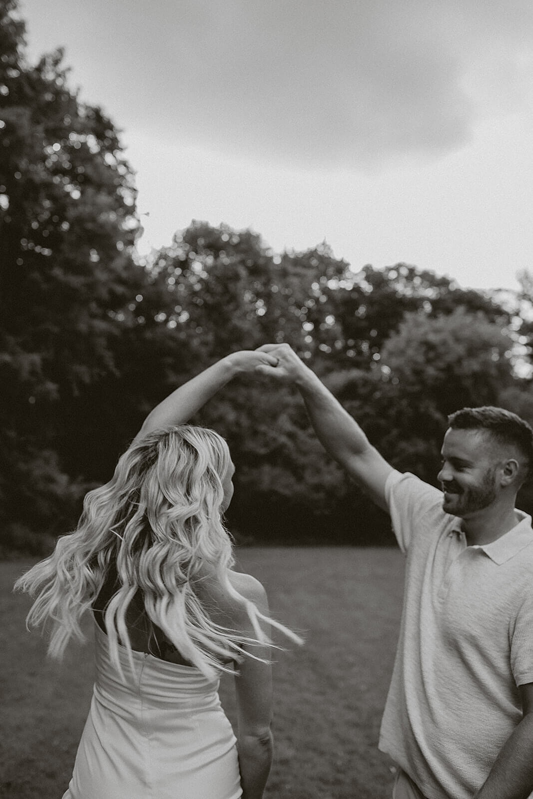 Black and white photo of a couple dancing together outdoors on a grassy field while holding hands beneath cloudy skies.