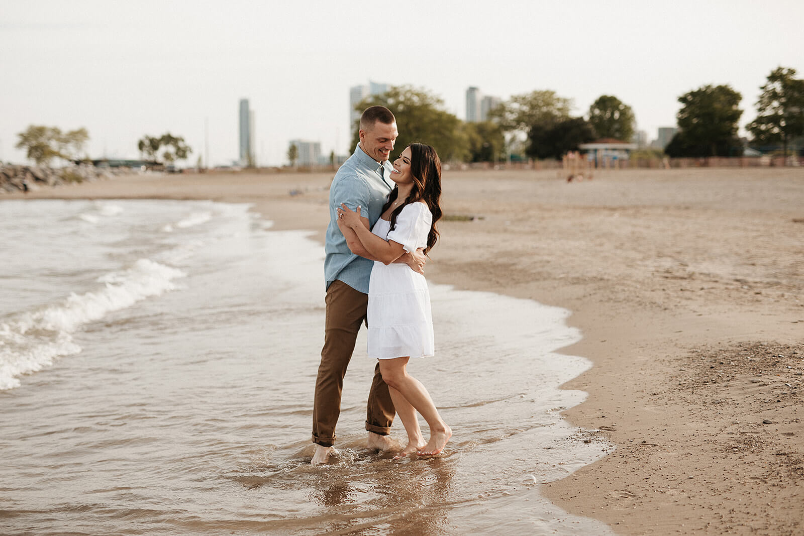 Couple embracing while standing barefoot at the edge of the beach with gentle waves washing onto the shoreline behind them.