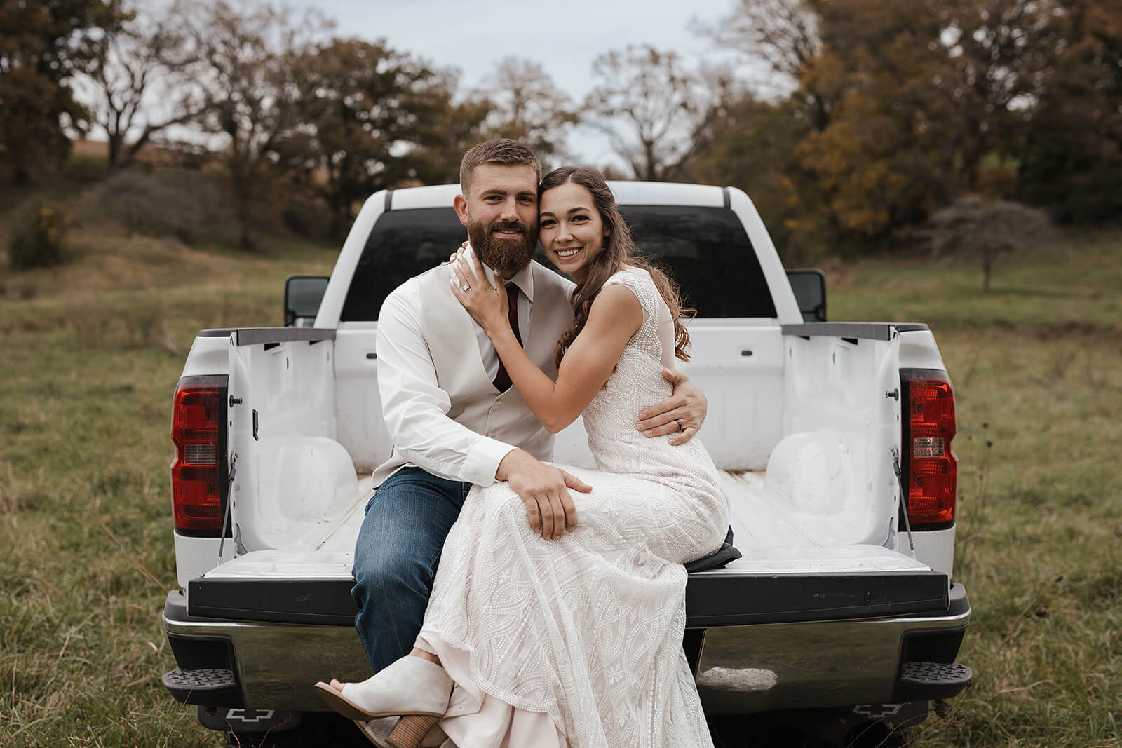 Bride and groom smiling together in the bed of a white pickup truck surrounded by fall trees during Lancaster Wisconsin wedding portraits