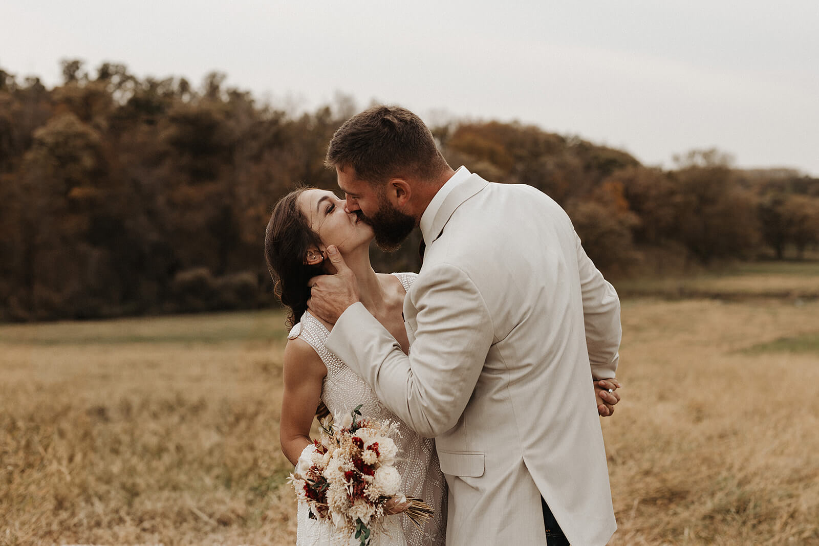 Bride and groom kissing in a golden field while she holds a loose bouquet of white and burgundy flowers during Lancaster Wisconsin fall wedding portraits