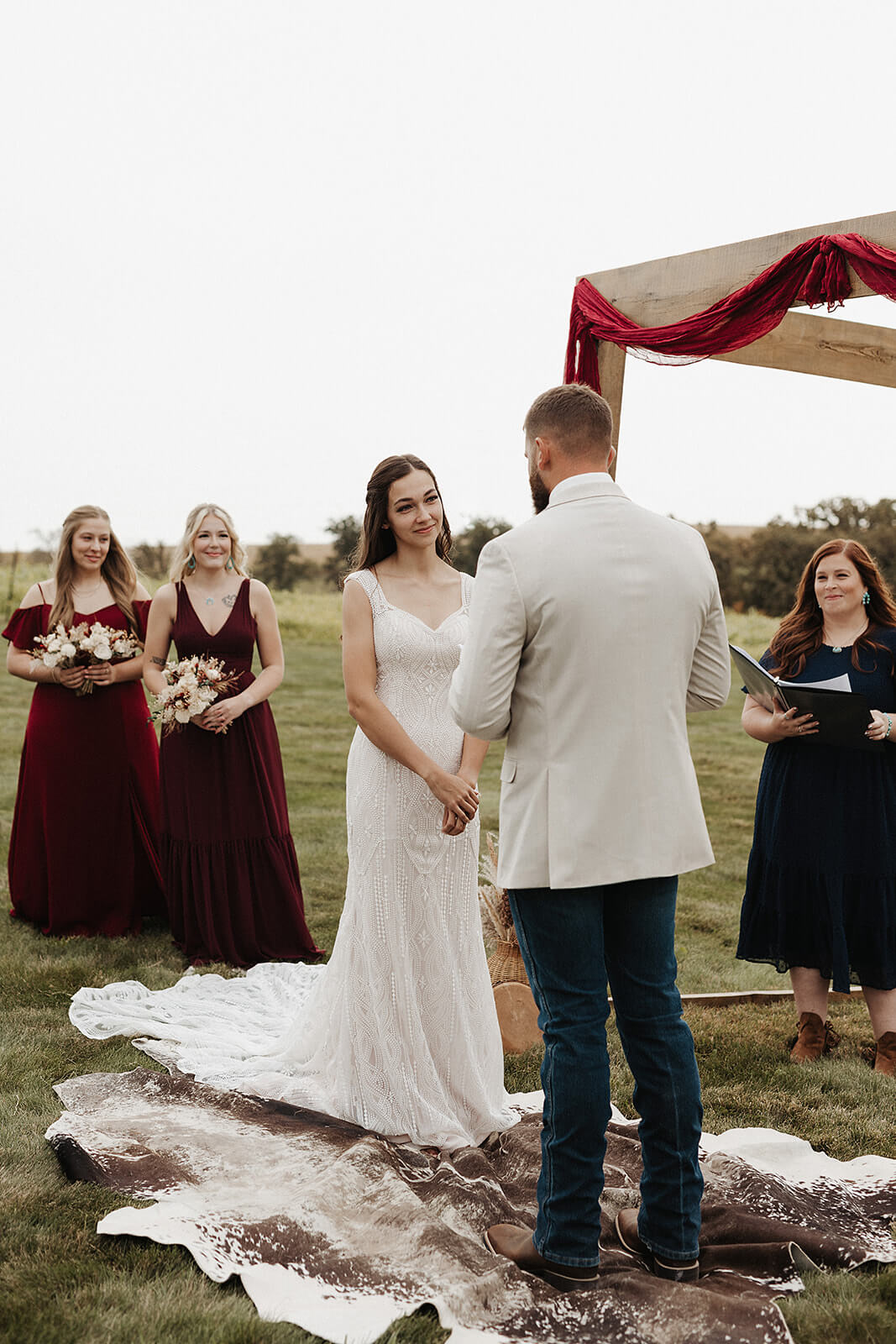 Bride and groom exchanging vows under a rustic wooden arch with bridesmaids in burgundy dresses at an outdoor Lancaster Wisconsin farmhouse wedding