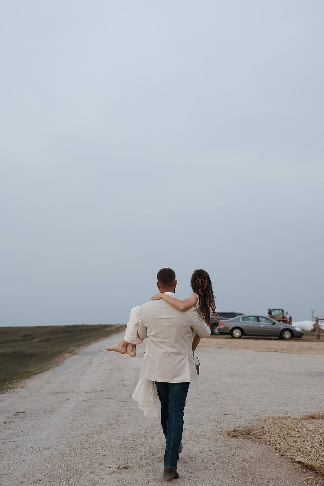 Groom carrying his bride along a gravel farm road during portraits at their Lancaster Wisconsin farmhouse wedding