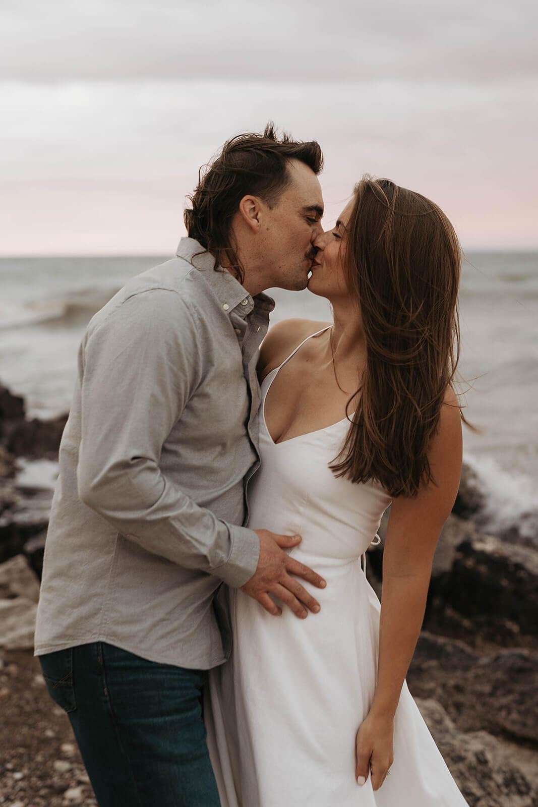 Couple kissing beside a rocky shoreline with waves crashing behind them during sunset on the beach.