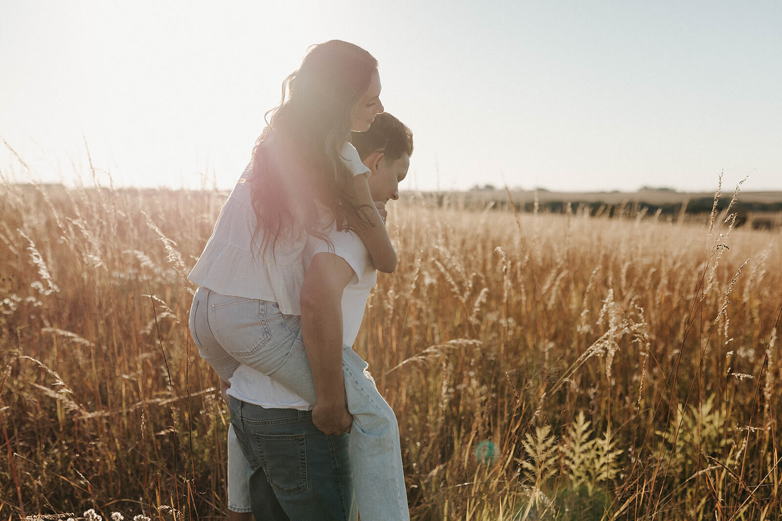 Woman riding piggyback on her partner’s back while walking through a golden grassy field during sunset.