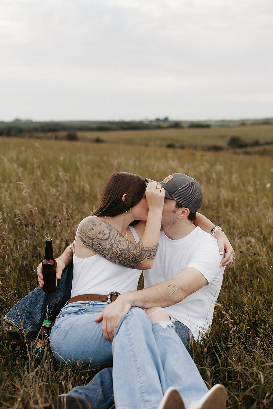 Couple sitting closely together in a grassy field during an outdoor engagement session, embracing while surrounded by tall golden grass and open countryside in the background.