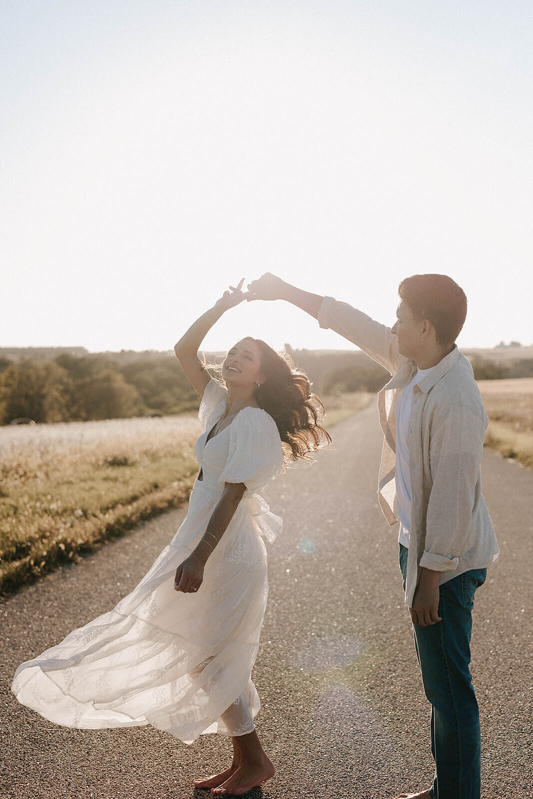 Couple dancing together on a country road during sunset with the woman’s white dress flowing in the wind and sunlight behind them.