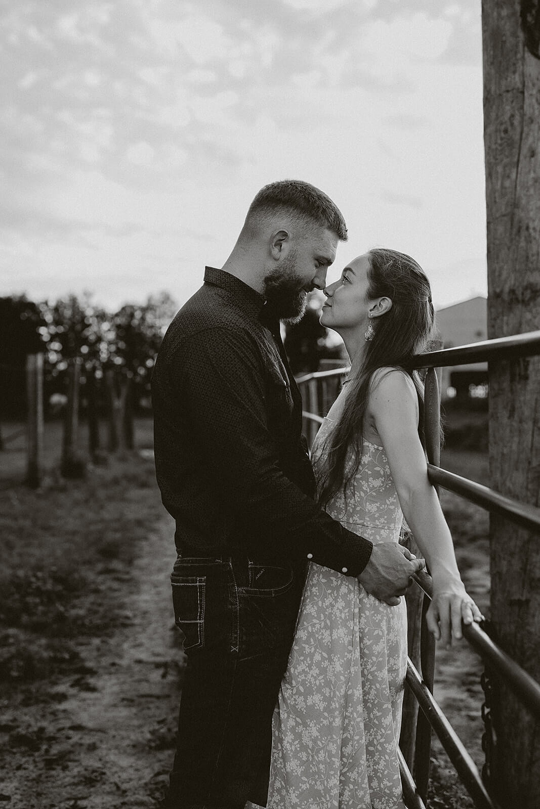 Black and white photo of a couple embracing beside a wooden fence during an outdoor engagement session in a grassy field.