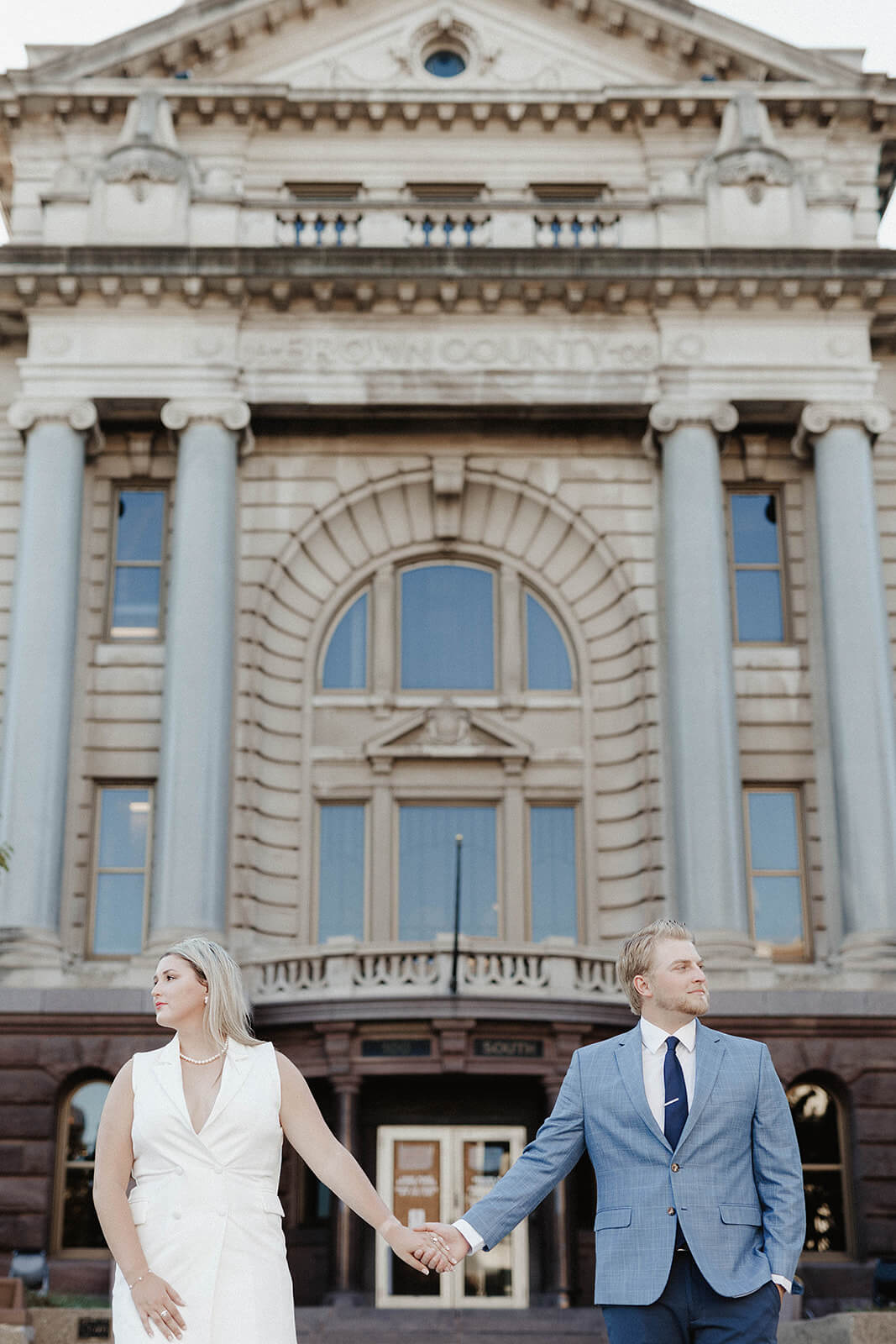 Couple holding hands in front of a large historic courthouse building with tall columns and ornate architecture.
