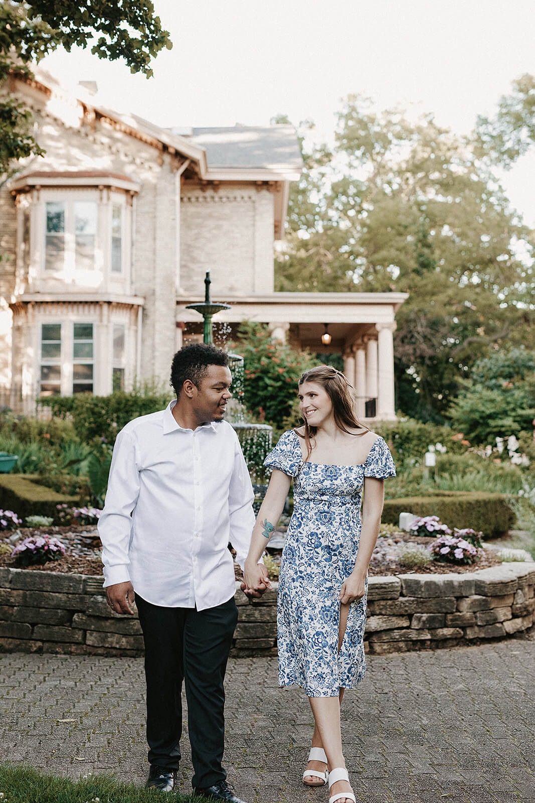 Couple walking together in front of a historic home and fountain during an outdoor engagement session with landscaped gardens in the background.