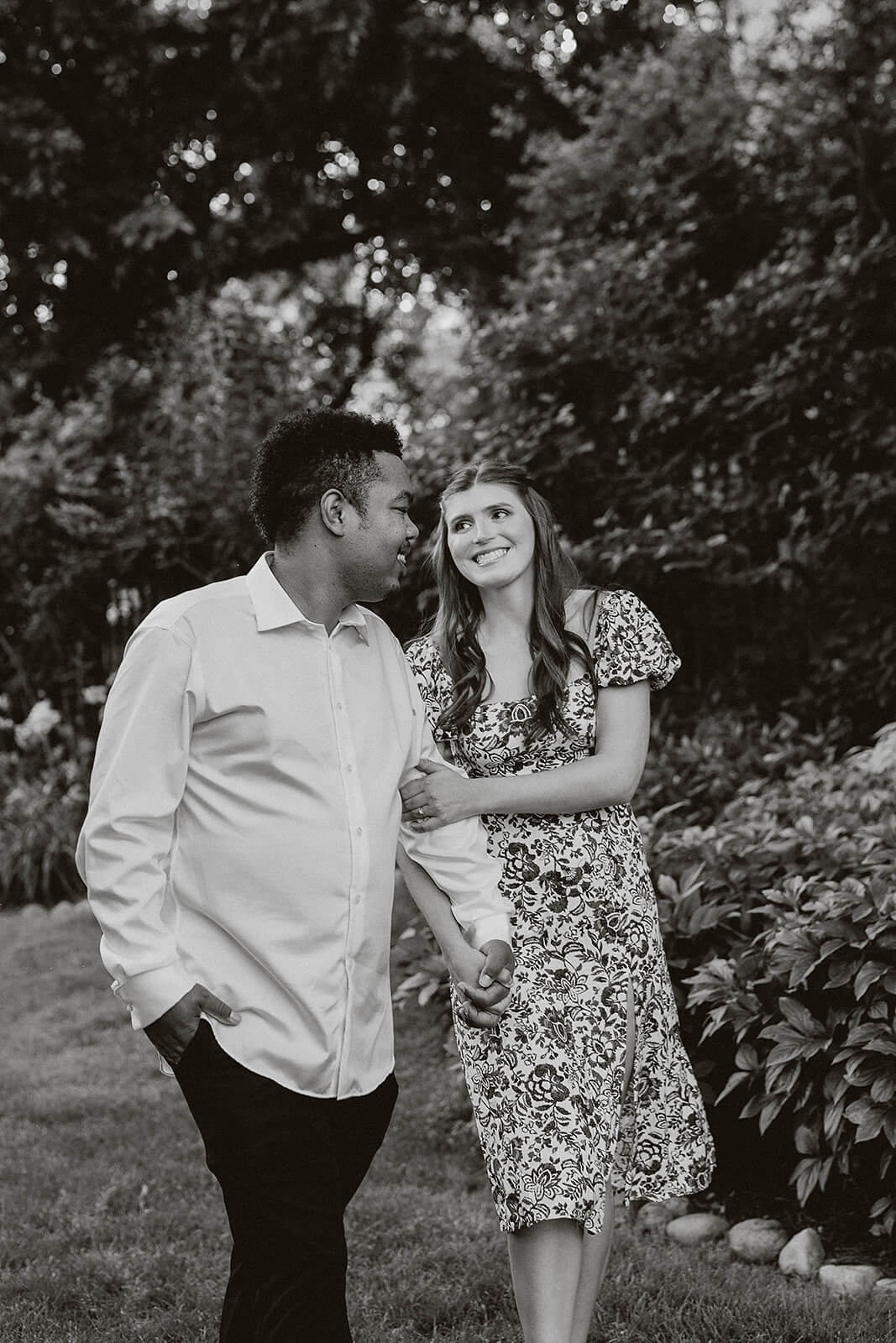 Black and white photo of a couple walking together through a garden path surrounded by trees and landscaping while holding hands and smiling at each other.
