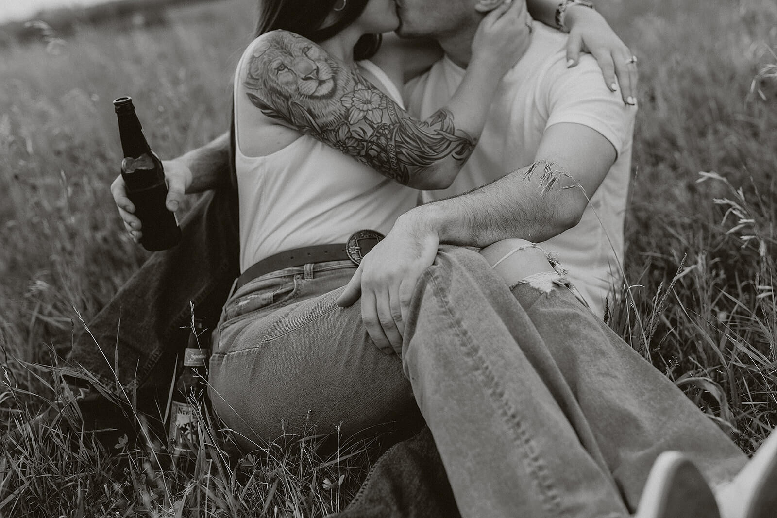 Black and white close-up photo of a couple embracing and kissing while sitting in a grassy field, with the woman holding a bottle and wearing a detailed floral tattoo sleeve.