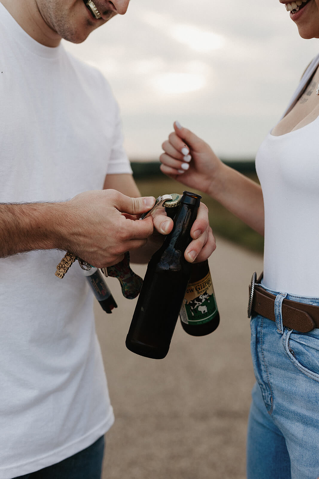 Close-up photo of a couple opening a bottle while standing on a gravel road during an outdoor engagement session, wearing casual white tops and denim jeans.