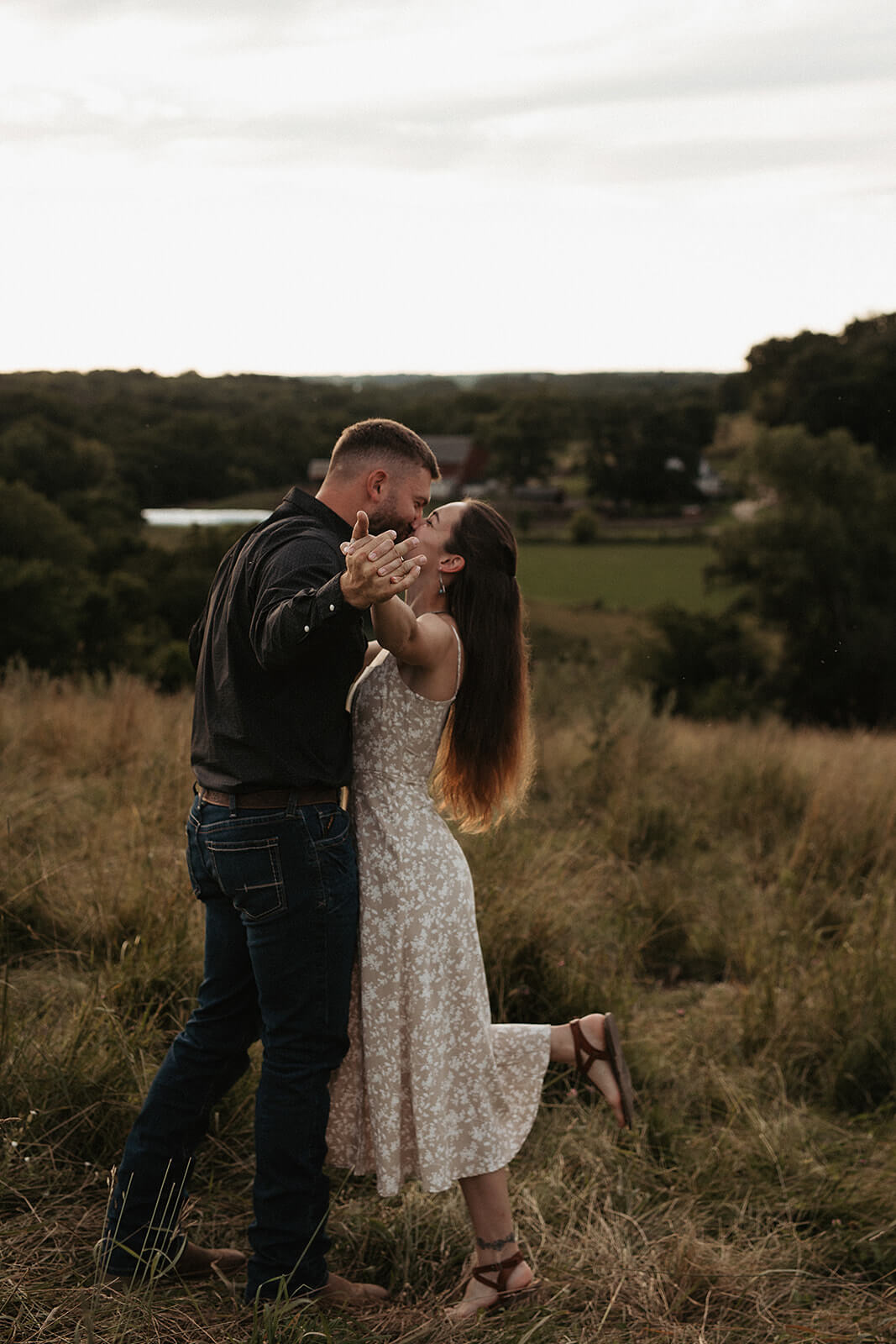 Couple dancing together in a grassy field during golden hour with rolling countryside and trees in the background.