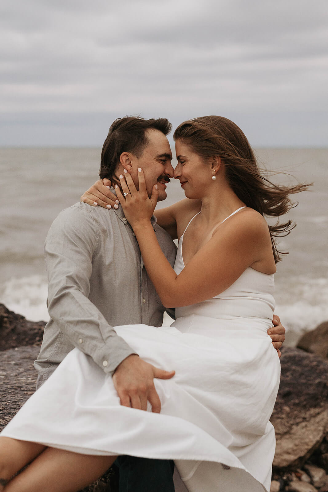 Couple sitting together on rocky shoreline cliffs beside the water while embracing during a beach engagement session on a cloudy evening.