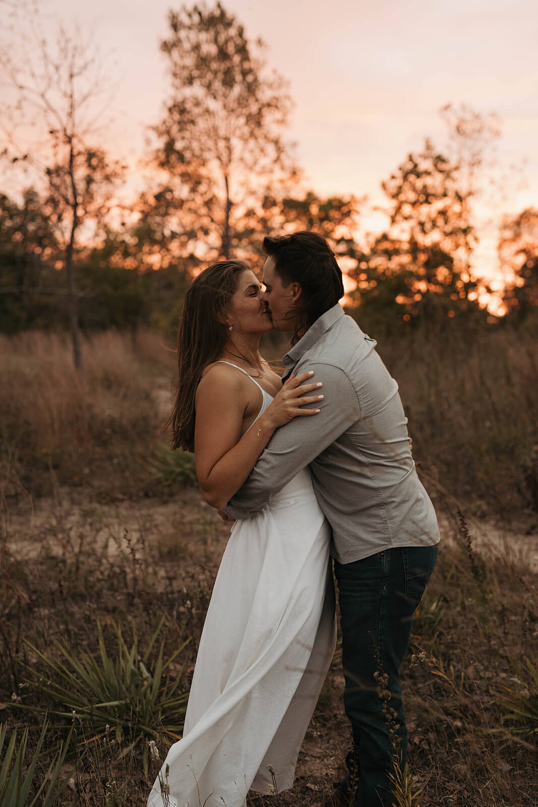 Couple embracing and kissing outdoors during sunset with warm golden light and tall grass surrounding them.