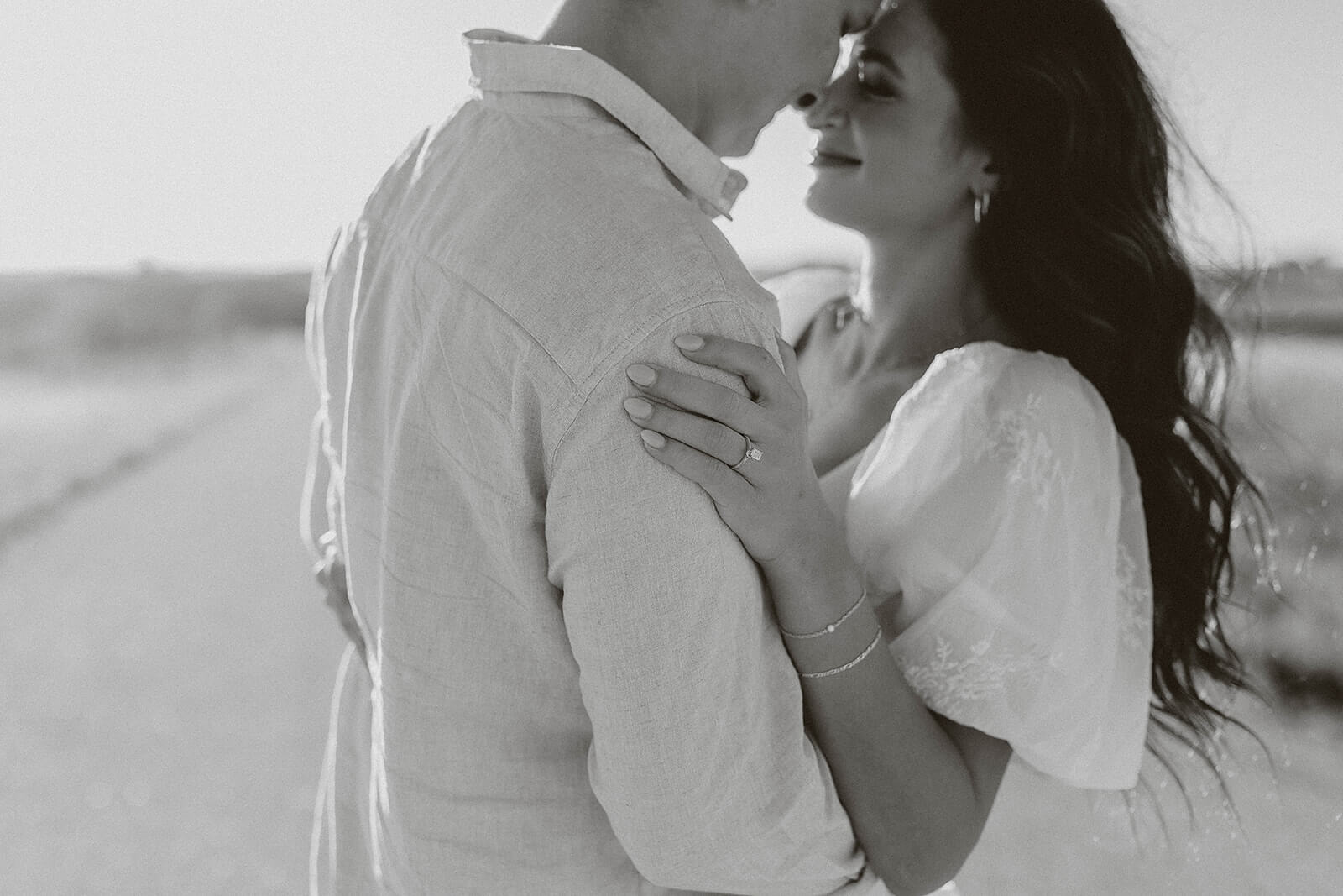 Black and white close-up portrait of a couple embracing outdoors during sunset, with the woman resting her hand on the man’s shoulder.