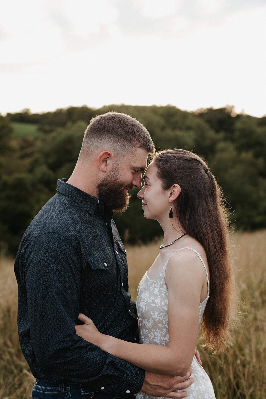 Close-up portrait of a couple standing forehead to forehead in a grassy field during golden hour with trees and rolling hills in the background.