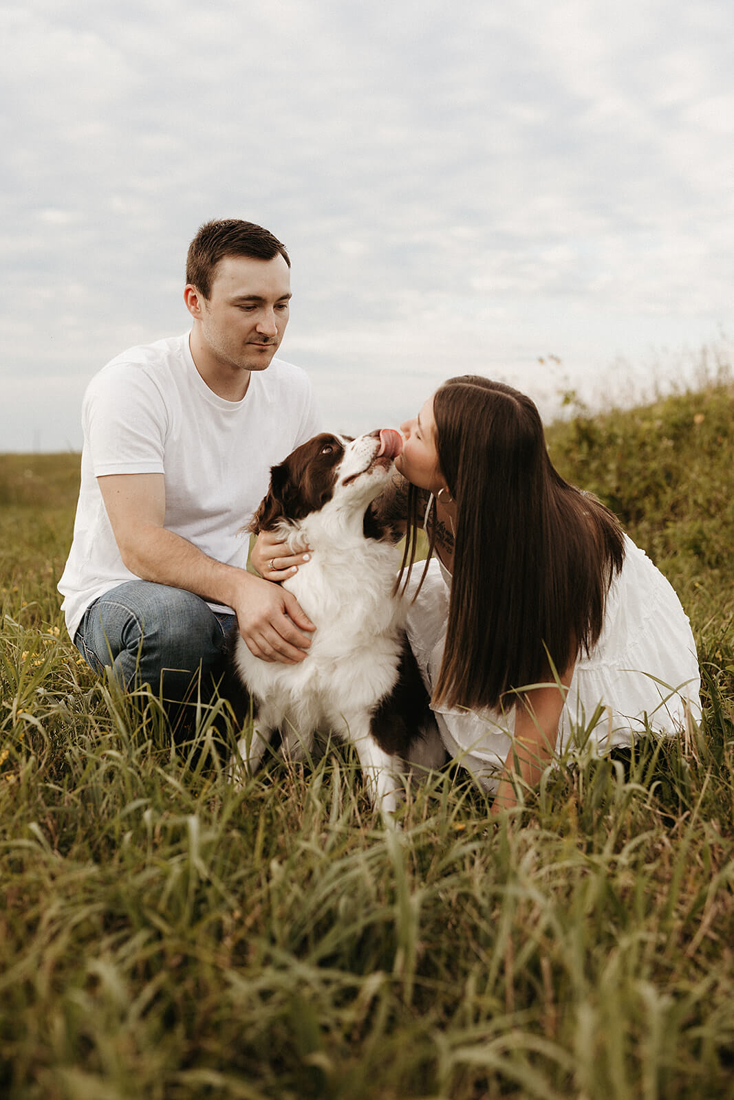 Couple sitting together in tall grass with their dog during an outdoor engagement session, surrounded by rolling hills and cloudy skies in the background.