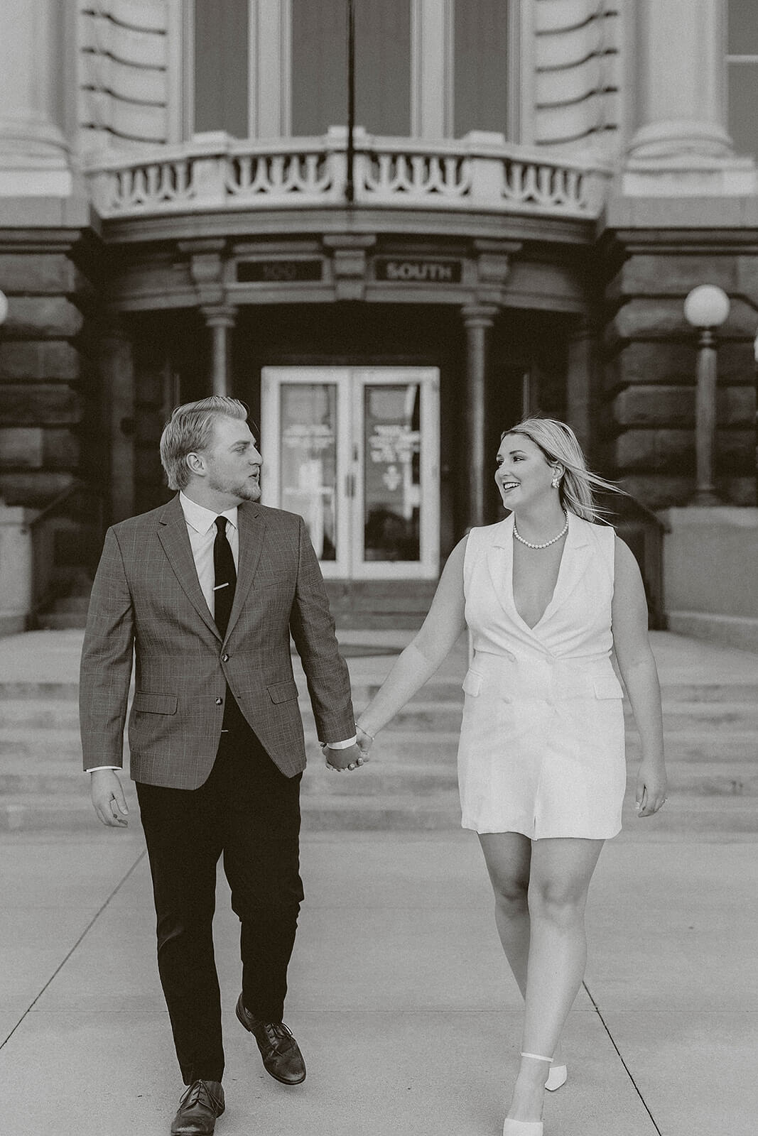 Black and white photo of a couple walking hand in hand in front of a historic courthouse entrance while smiling at each other.