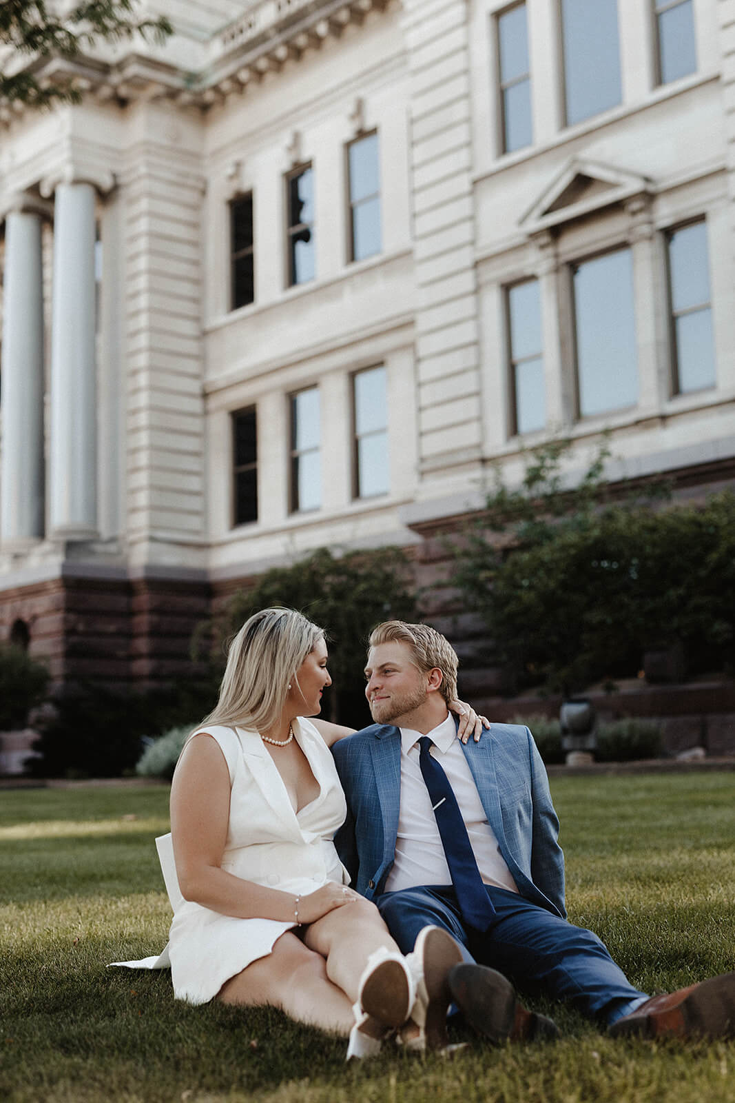 Couple sitting together on the grass in front of a historic courthouse building during an elegant downtown engagement session.