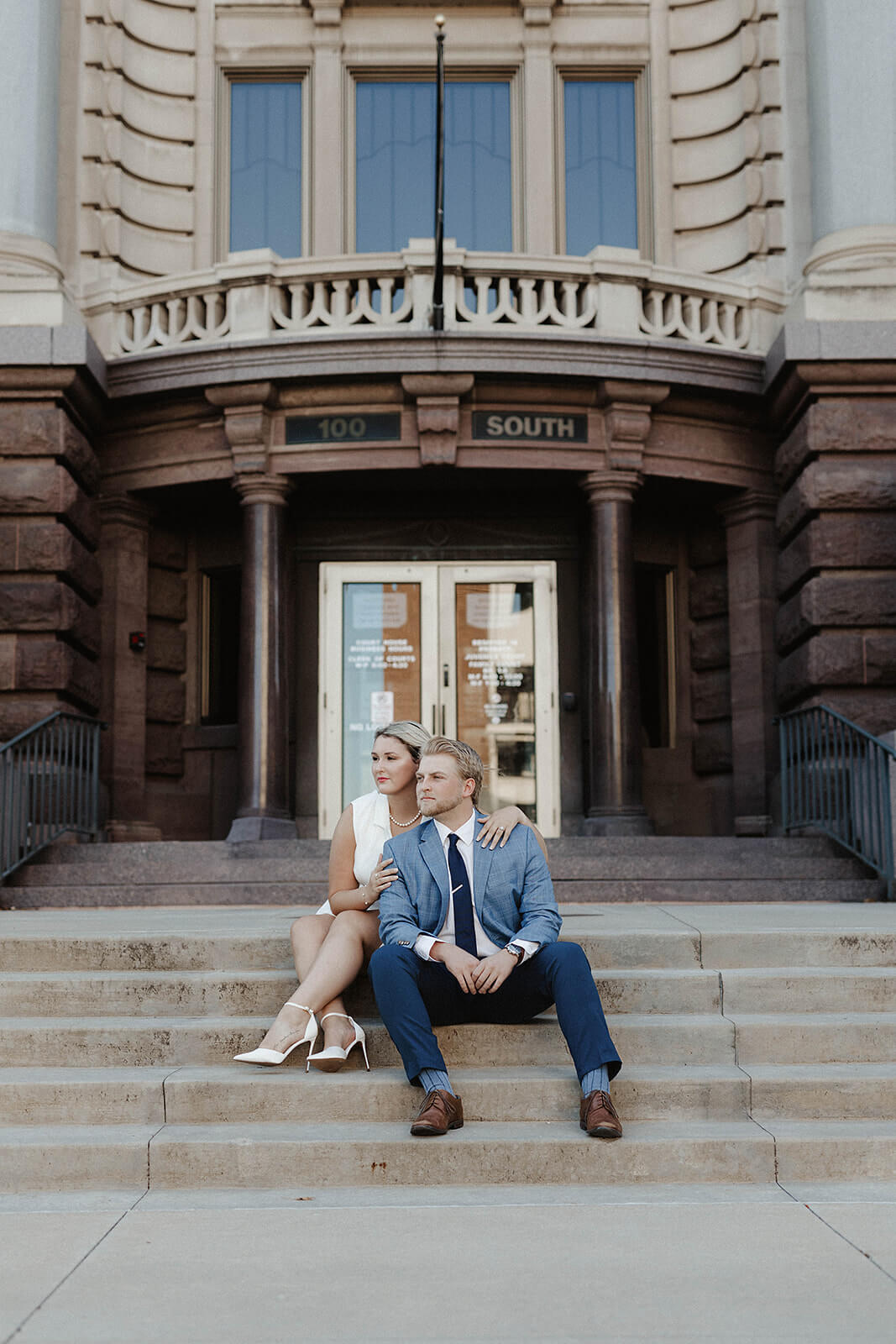 Couple sitting together on the stone steps in front of a historic downtown building with ornate architecture and large wooden doors during a city engagement session.
