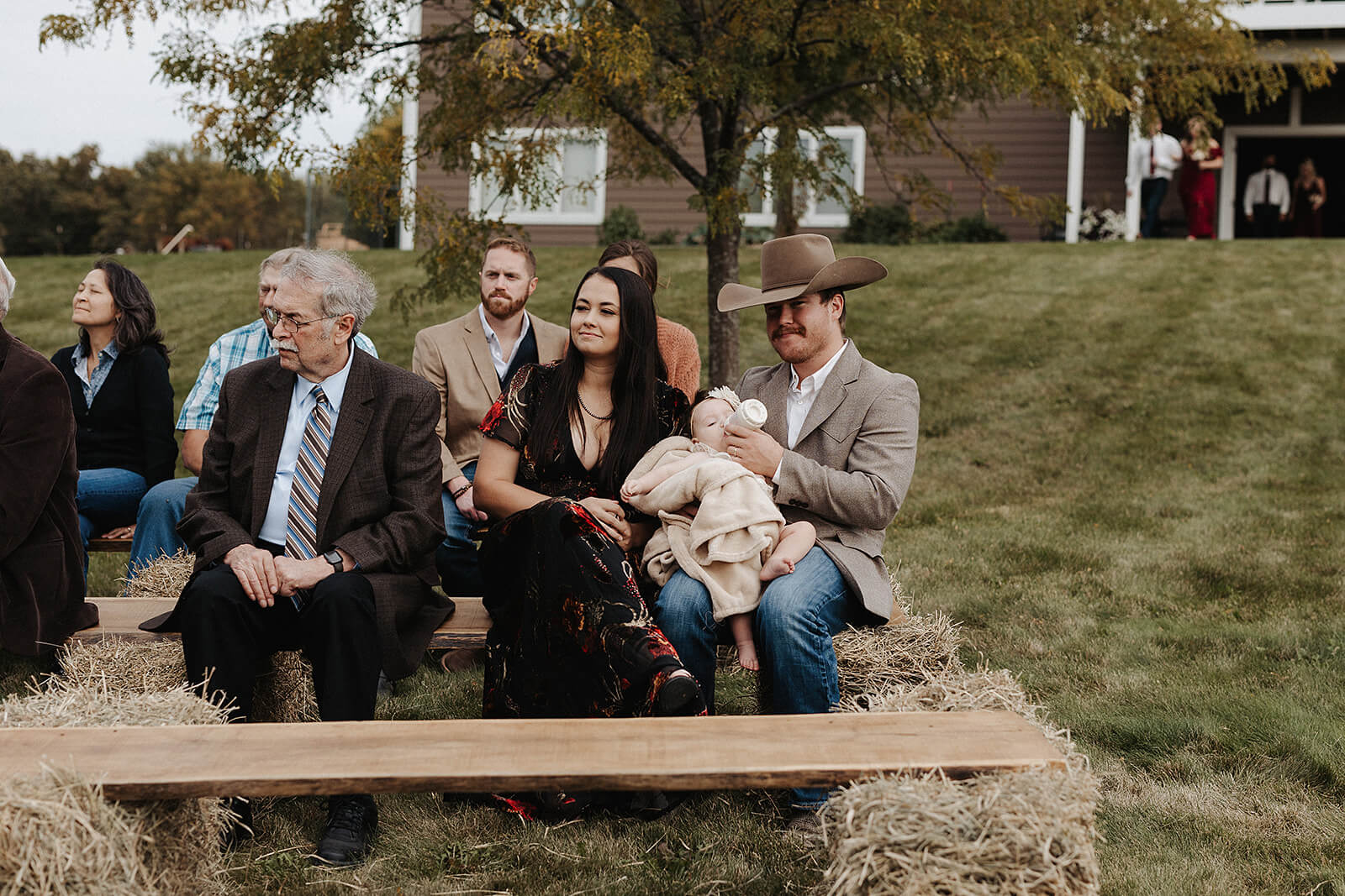 Wedding guests seated on wooden benches and hay bales watching the outdoor ceremony at a Lancaster Wisconsin farmhouse wedding