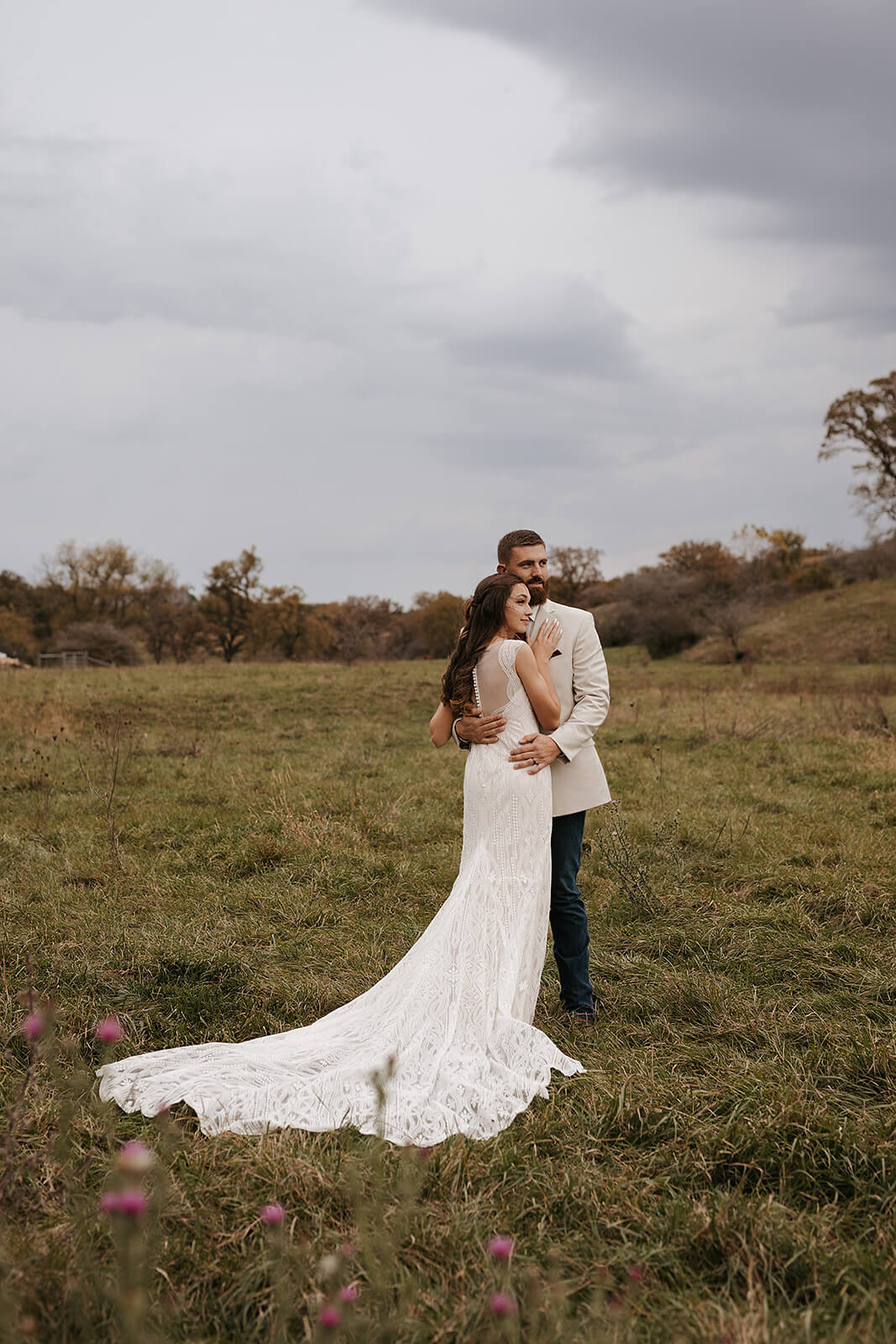 Bride and groom posing together in a green field with a dramatic cloudy sky and lace wedding gown train trailing behind them at a Lancaster Wisconsin farmhouse wedding
