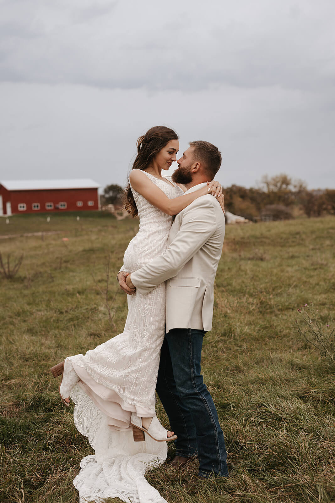 Bride and groom embracing in front of a red barn with lace wedding dress train trailing across the grass at a Lancaster Wisconsin farmhouse wedding