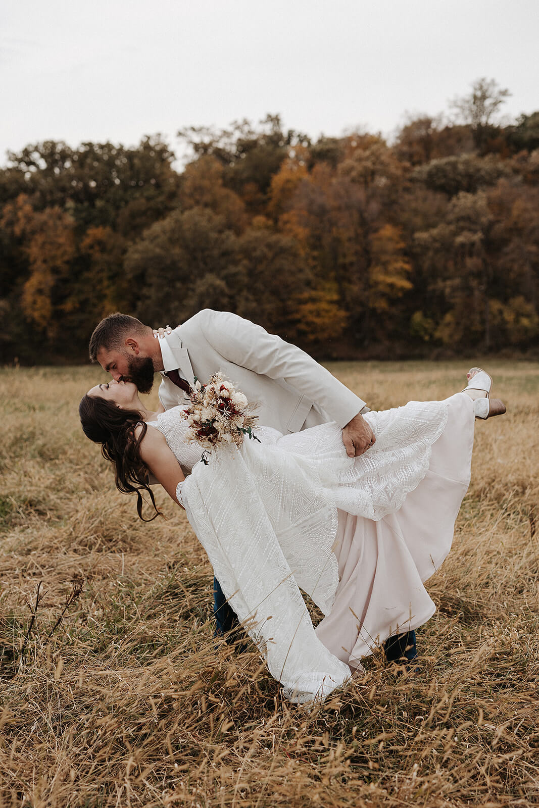 Groom dipping his bride for a kiss while holding her bouquet during fall wedding portraits in a golden field at a Lancaster Wisconsin farm