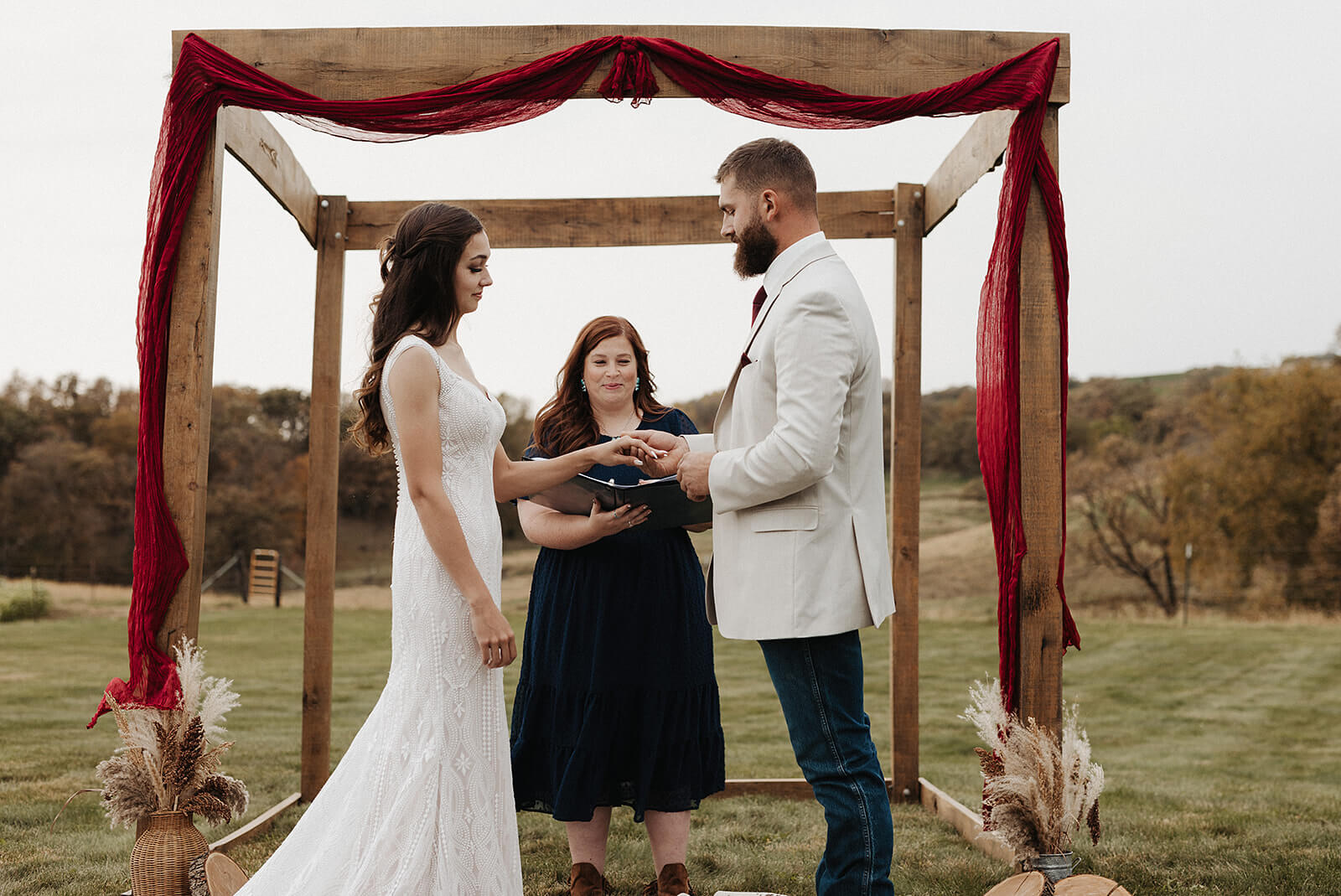 Bride and groom exchanging rings under a rustic wooden arch draped in red fabric during their outdoor Lancaster Wisconsin farmhouse wedding ceremony