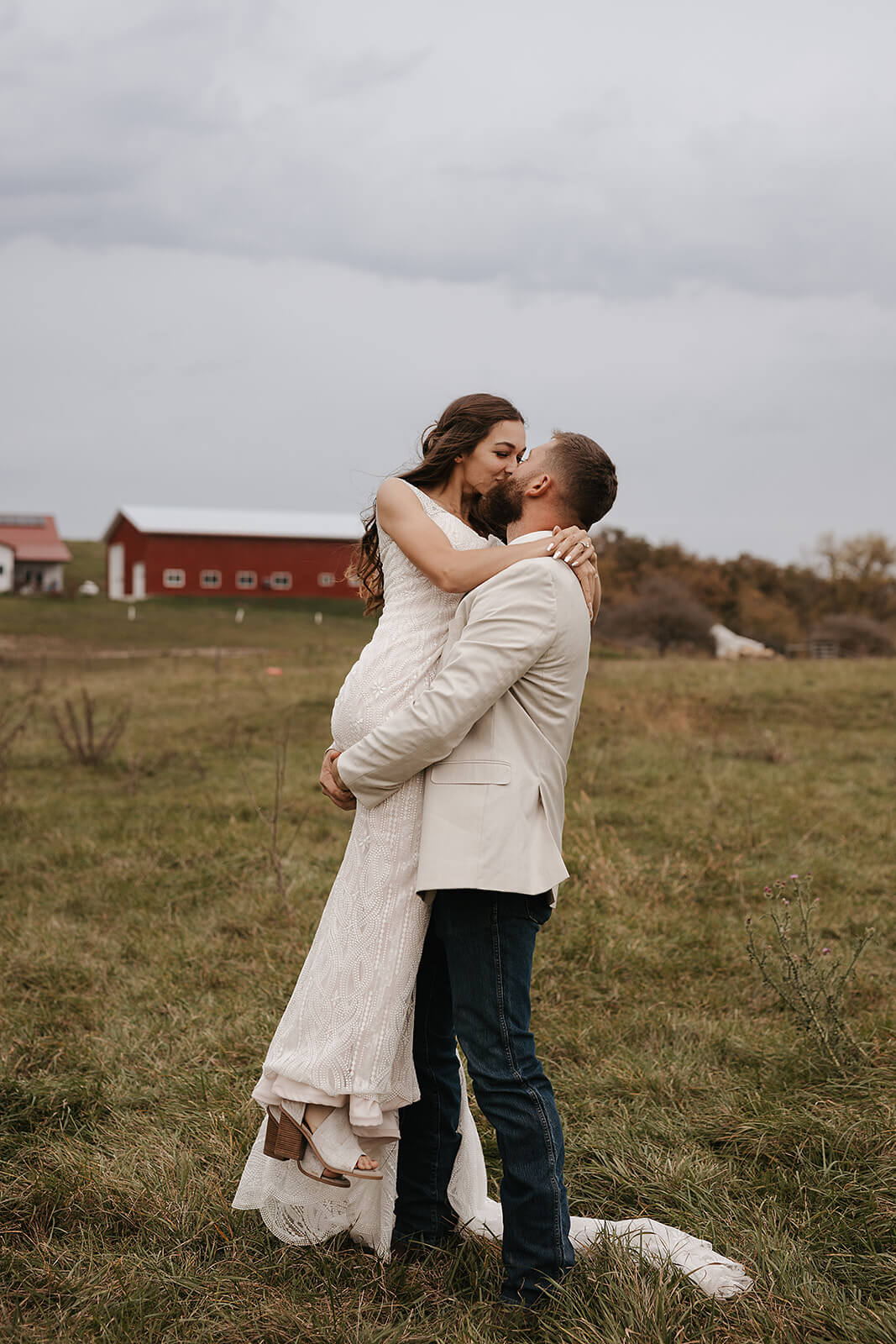 Bride and groom kissing in a field with a red barn visible on the hill behind them at a Lancaster Wisconsin farmhouse wedding